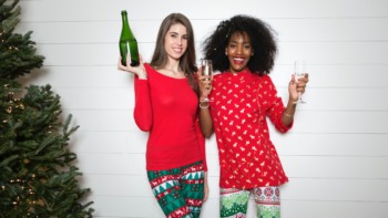 two women pose with a Christmas tree and champagne.
