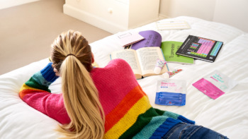 Female student lays on her bed with books and superdrug essentials surrounding her.