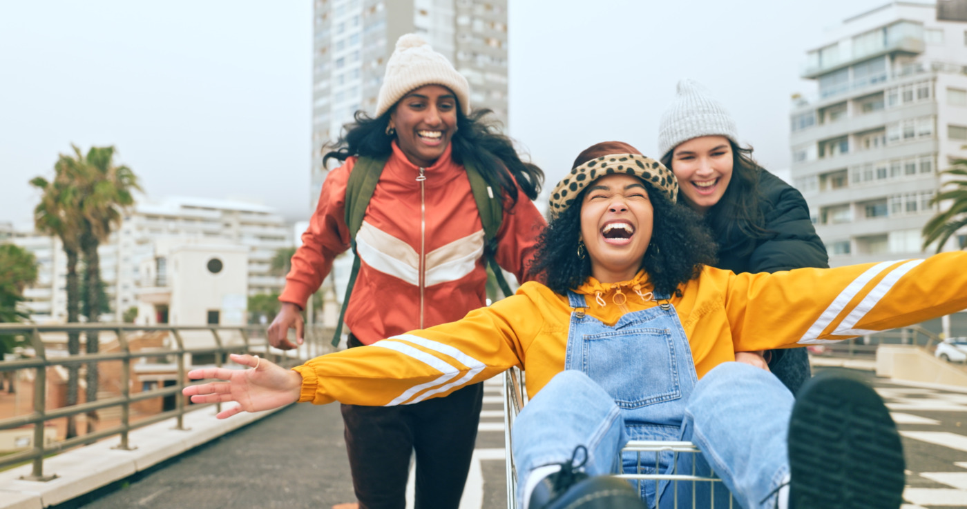 Three Gen Z woman laughing together. One is being pushed in a shopping trolley. 