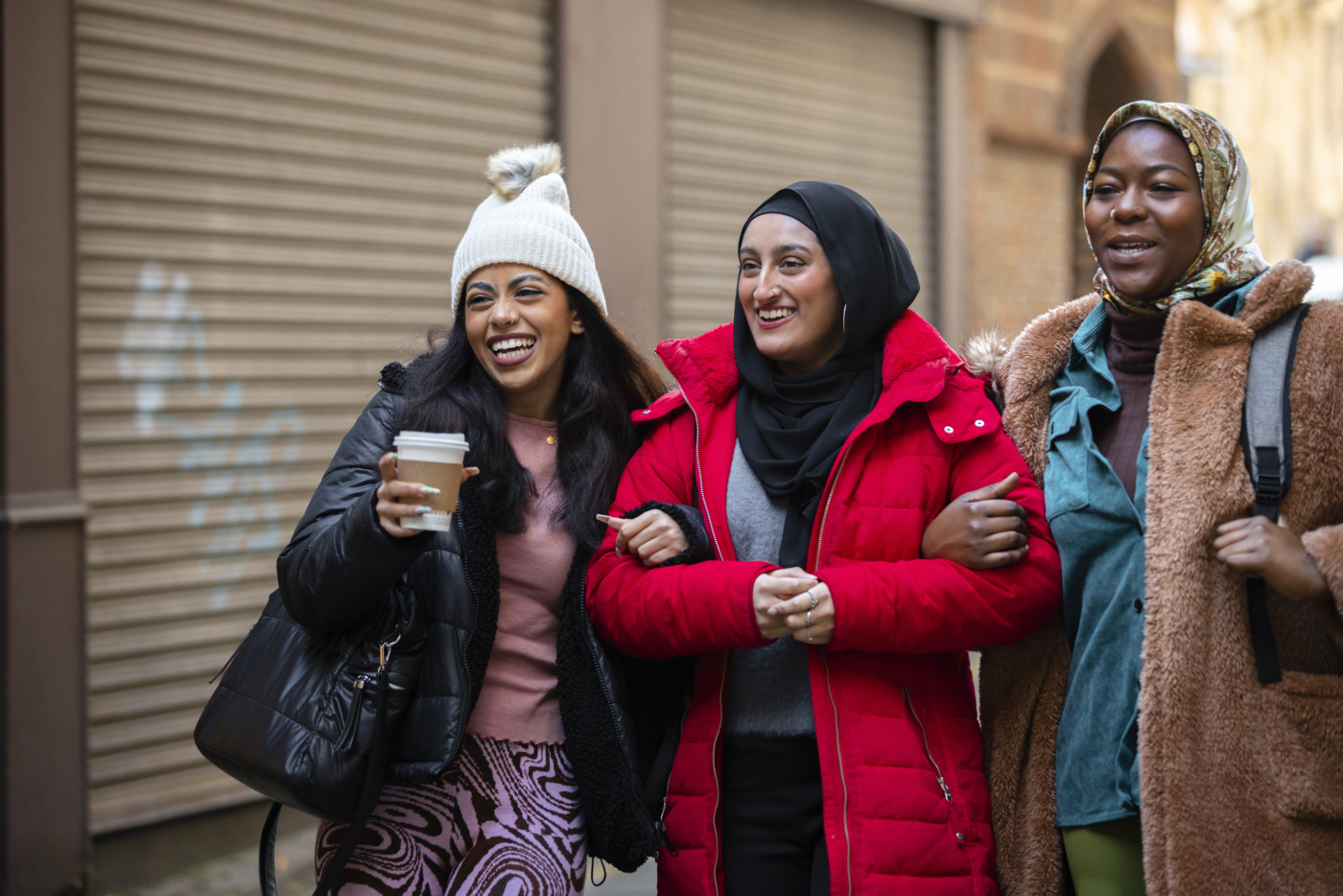 A medium shot of three young women wearing warm casual clothing,  in Newcastle Upon Tyne. They are walking down an alley in the city linked in with each other, talking and having a laugh.